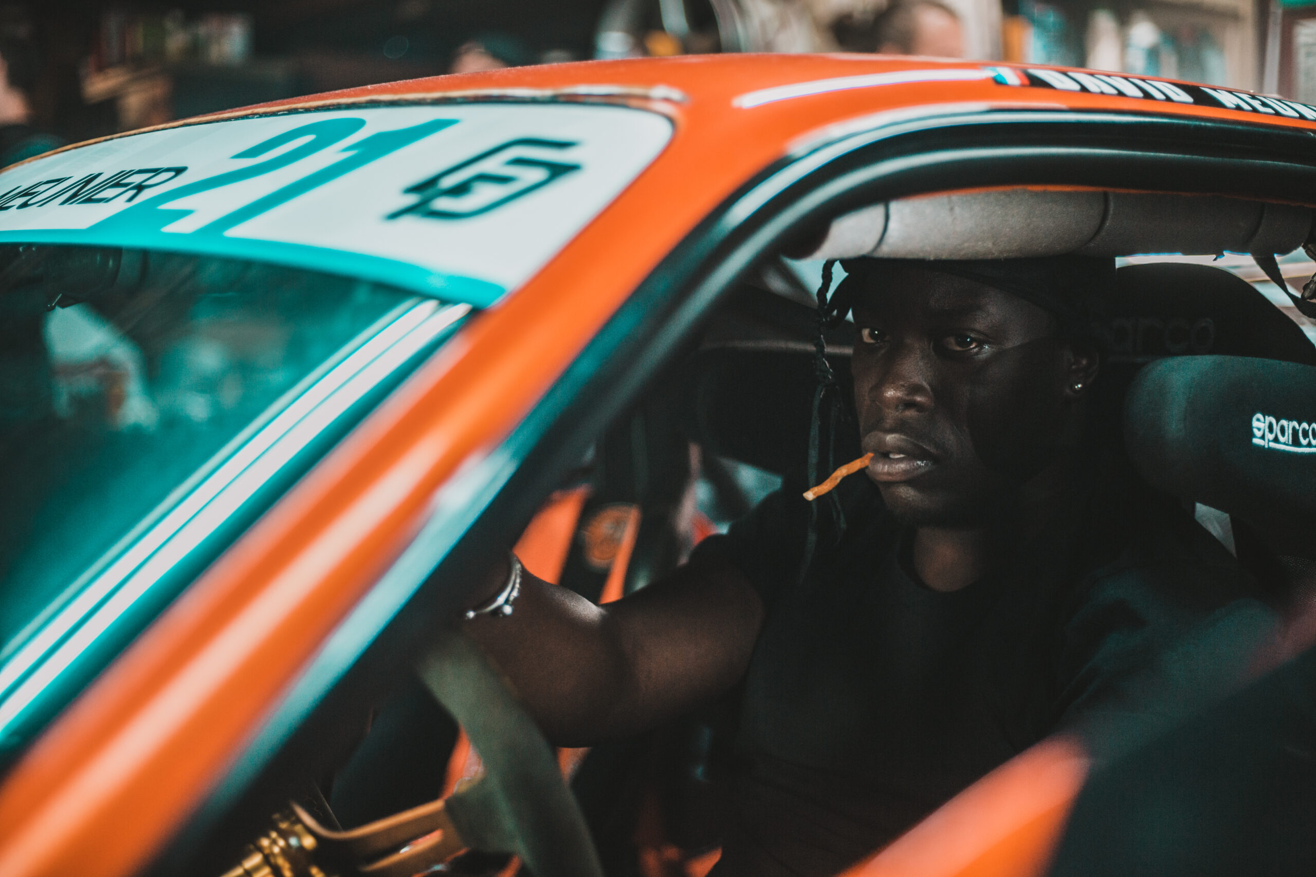 un close-up d'un rappeur dans une voiture orange, qui regarde le caméra , cigarette en bouche